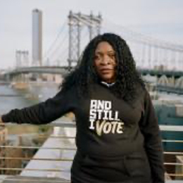 Rasheta Bunting is standing in front of the manahttan bridge posed with her arm laying over a silver banister. She has black curly hair and is wearing a black hoodie that says “And still I vote”.